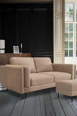 The Grace Medium Sofa In Luxe Needlecord Velvet Natural Whisper styled in a dark living room with the matching footstool, with a sideboard and some home accessories in the background.