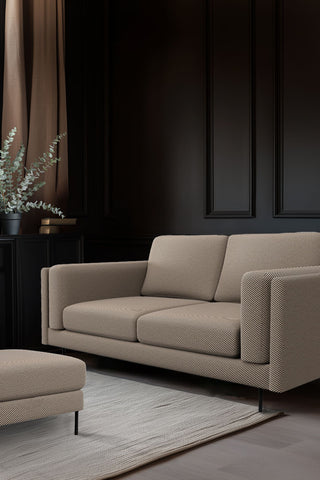 The Grace Medium Sofa In Studio Stripe Velvet Sand & Black styled in a dark living room with the matching  footstool, with a rug, some books and a potted plant.