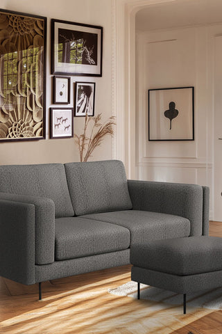 The Grace Medium Sofa In Timeless Texture Black Bean styled in a living room with the matching footstool, with various art prints on the wall and some pampas grass behind the sofa.