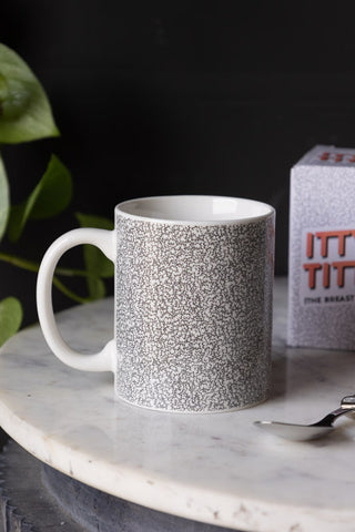 The Itty Bitty Titty Mug displayed on a marble table with the box and a spoon, with a plant in the background.