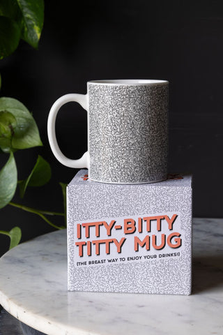 The Itty Bitty Titty Mug displayed on top of the box, on a marble table with a plant in the background.