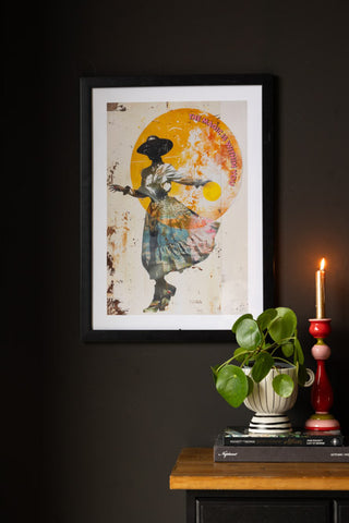 The Magic Is Within You Art Print styled on a black wall, above a sideboard with books, a planter and a lit candle in a colourful candlestick holder.