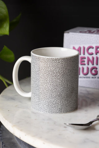 The Micro Penis Mug displayed on a marble table with the box and a spoon, with a plant in the background.