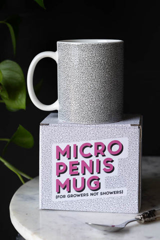 The Micro Penis Mug displayed on a table on the box, next to a spoon, with a plant in the background.