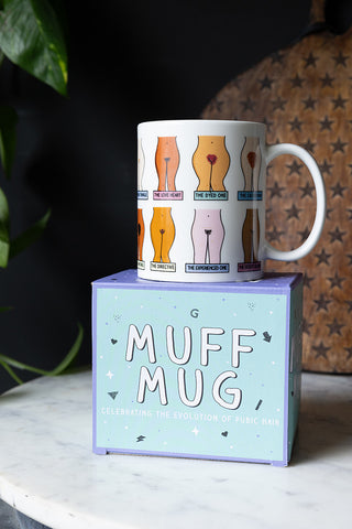 The Muff Mug styled on its box, displayed on a marble table in front of a wooden serving board and a plant.