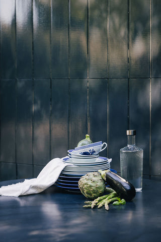 The Paveau - Gorgeous Ribbed Glass Carafe in White styled on a table with vegetables, a large stack of plates and a tea towel.