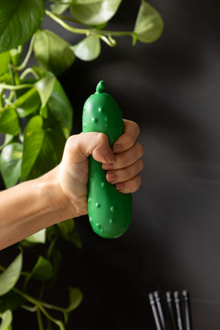 The Stress Pickle being squeezed by a hand, in front of a dark background with a plant.