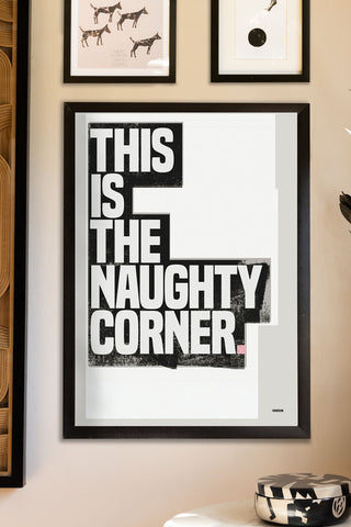 The This Is The Naughty Corner Monochrome Art Print - Unframed styled in a black frame, displayed on a wall with other art prints, above a table with a trinket box.