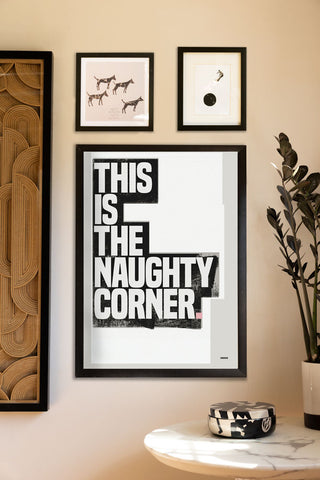 The This Is The Naughty Corner Monochrome Art Print - Unframed styled on a wall in a black frame, with other art prints and a table with a trinket box and a potted plant.