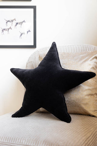 The Velvet Star Cushion In Black displayed on a chair with another cushion, with an art print on the wall in the background.