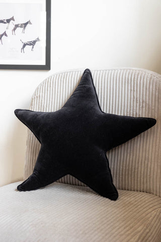 The Velvet Star Cushion In Black styled on a neutral chair, with a framed art print on the wall in the background.