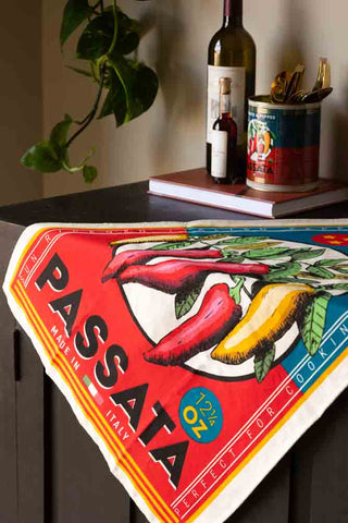 The Vintage Passata Tea Towel displayed draped over the edge of a wooden sideboard, with a book, some bottles, a passata storage tin containing cutlery and a plant in the background.