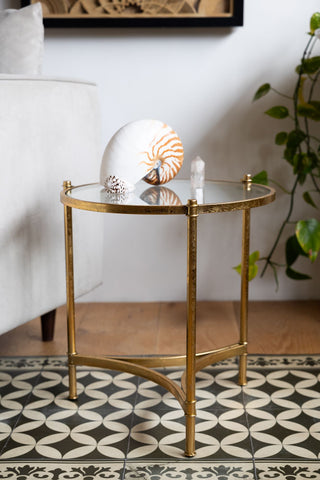 The Vintage-Style Gold & Glass Side Table displayed next to a chair in a living room, styled with some ornaments and a plant.