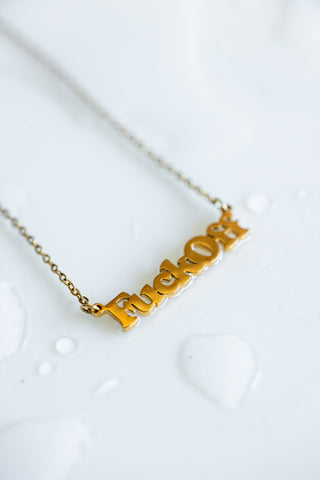 The Waterproof Fuck Off Gold Necklace on a white surface with water droplets.