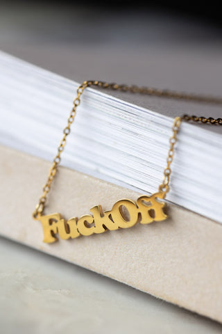 The Waterproof Fuck Off Gold Necklace draped over the corner of a neutral surface.