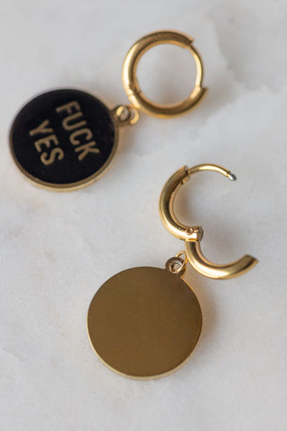 The Waterproof Fuck Yes Gold Charm Earrings displayed together on a neutral surface, with one face down with the clasp open.