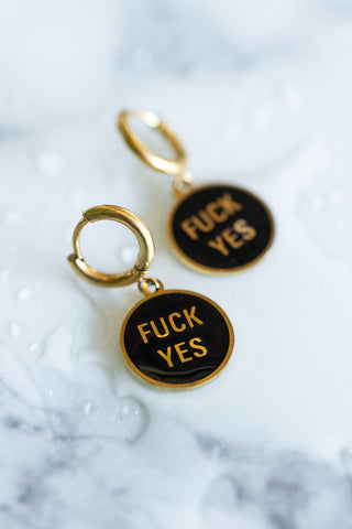 The Waterproof Fuck Yes Gold Charm Earrings displayed on a marble surface with water splashes.