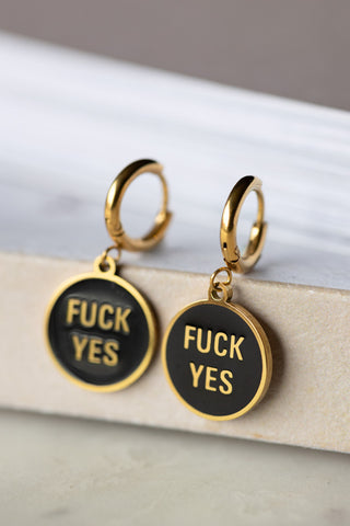 The Waterproof Fuck Yes Gold Charm Earrings displayed on the corner of a neutral surface.