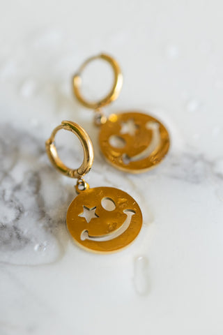 The Waterproof Gold Wink Face Charm Earrings displayed on a white marble surface with water drips.