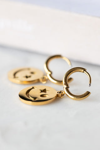 The Waterproof Gold Wink Face Charm Earrings displayed on a white surface with the clasps open.