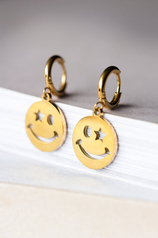 The Waterproof Gold Wink Face Charm Earrings displayed on a white surface.