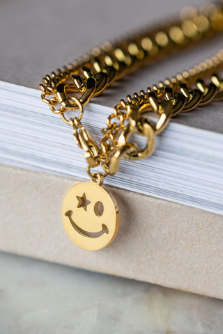 Th Waterproof Wink Face Gold Chain Bracelet draped on the corner of a neutral surface, with the charm hanging.