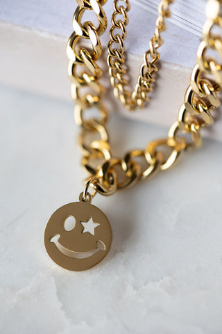 The Waterproof Wink Face Gold Duo-Chain Necklace on a white marble surface.