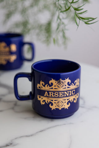 The What's Your Poison - Arsenic Mug shown on a white marble surface with a green plant in the background.