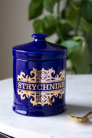 the What's Your Poison - Strychnine Jar displayed on a white surface with a gold spoon and a plant in the background.