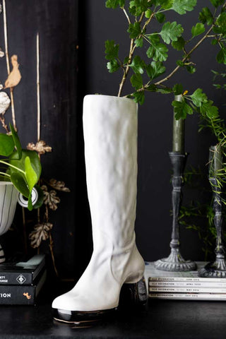 The Fabulous White Boot Vase styled with greenery inside, displayed on a black surface with other home accessories.