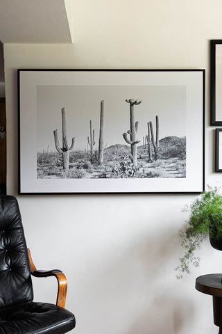 The Wild West Cactus Art Print displayed on a white wall with some other prints, styled with a chair, side table and a potted plant.