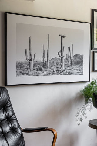 The Wild West Cactus Art Print displayed on a white wall with other art prints, styled above a chair, side table and a plant in a pot.