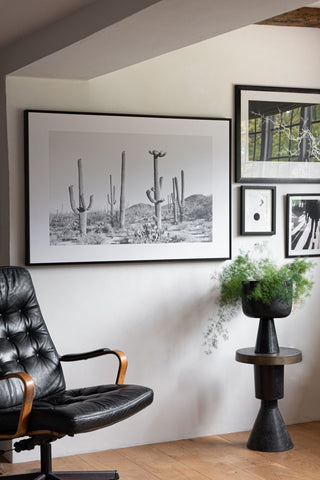 The Wild West Cactus Art Print styled on a wall with other art prints, displayed above a side table with plant and a desk chair.