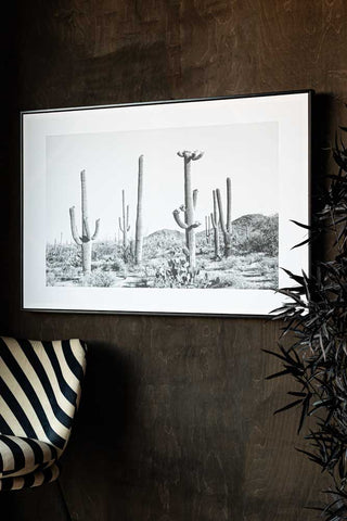 The Wild West Cactus Art Print displayed on a dark wooden tall above a stripy chair and a plant.