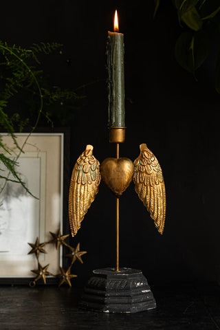 The Winged Milagro Heart Candle Holder with a lit candle inside, styled on a dark surface in front of a photo frame, star decoration and greenery.