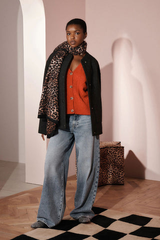 A model standing on a patterned rug, wearing the Woven Leopard Print Scarf, with a leopard print stool in the background of the shot.