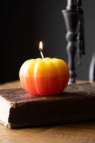 A lit pumpkin candle styled on a book on a wooden surface, with a candlestick with melted wax in the background.