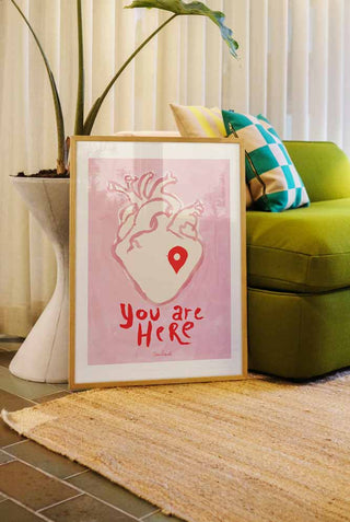 The You Are Here Art Print styled in a frame in a living room, leaning against a sofa and a plant.