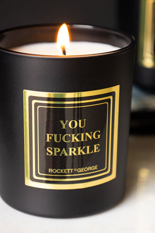 Detail shot of the design on the lit You Fucking Sparkle Scented Candle.