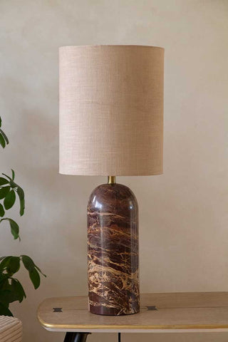 The Wine Red Marble Table Lamp with a shade on, styled on a wooden bench next to a plant.