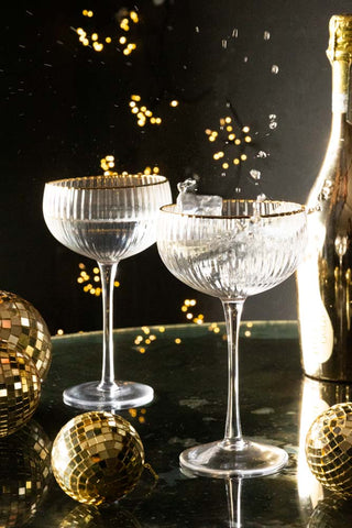 Ribbed coupe glasses with ice being dropped into the glass. In the background are small gold disco balls and fairy lights.