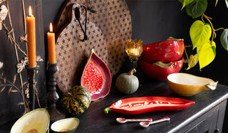 An autumnal lifestyle shot of various kitchenware items displayed together on a black sideboard with some lit candles and some greenery.