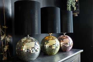 3 Disco ball lamps on a black sideboard in a kitchen. The disco balls are in silver, gold and pink with black linen shades.