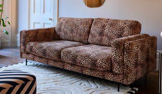 A leopard print sofa styled in a living room on a rug, with a mirror, plant and footstool.