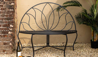 The Lotus Leaf Rounded Back Garden Bench outside in front of a neutral wall, styled with a plant, a wine bottle and serveware.