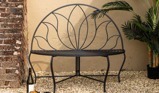 The Lotus Leaf Rounded Back Garden Bench styled in front of a neutral wall with a potted plant, wine bottle and serveware.