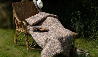 The Floral Paisley Bench Seat Pad displayed on a wicker lounge chair in a garden, styled with a hat, serving board with snacks and two lanterns on the floor.