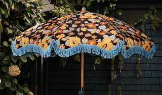 The HKliving Floral Flourish Parasol with Blue Fringing styled in a garden, in front of a black building with lots of greenery.