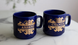 Two mugs with the words 'Arsenic' and 'Cyanide' written on them in a decorative design, displayed on a white marble table.