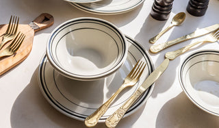 A dining table with plates, bowls, cutlery and various other dining accessories on, in the sunshine.